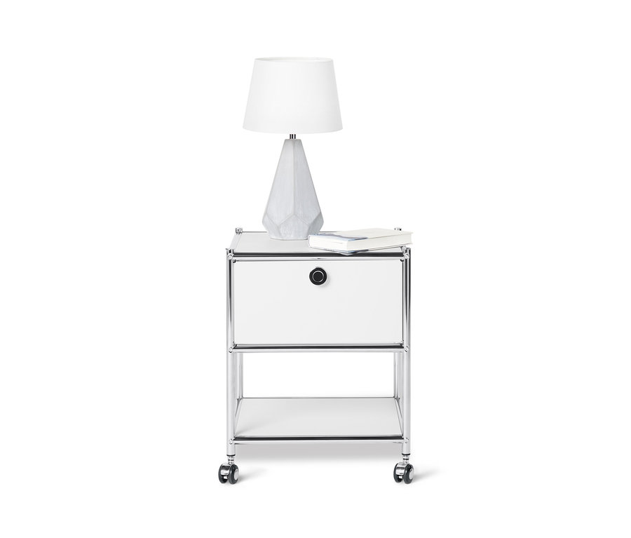 Sur un fond blanc, une table d'appoint en métal blanc avec tiroir « CN3 », sur laquelle reposent une lampe et un livre.