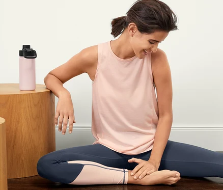 Une femme est assise par terre et porte 2 hauts de sport et des leggings.