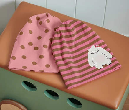 Deux bonnets roses et marrons sont posés sur un coffre à jouets marron-beige : un bonnet rose à pois marron et un bonnet rayé rose et marron avec un patch d’un monstre blanc.