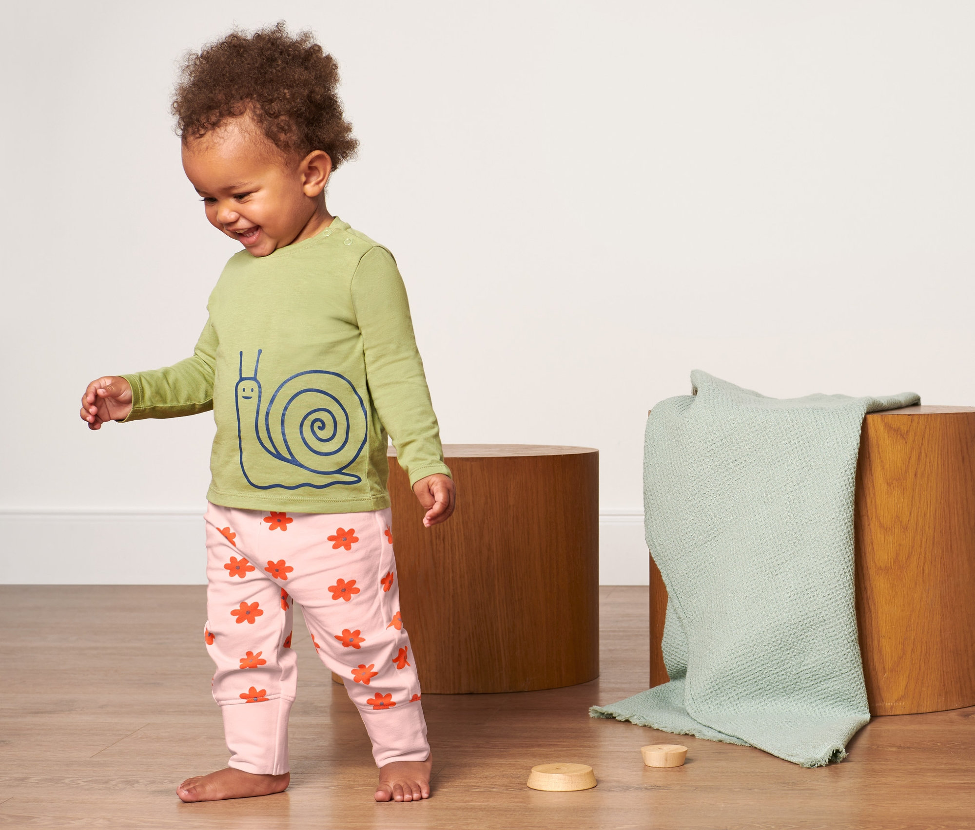 Un bébé souriant se tient sur le sol et porte un pantalon de survêtement rose à motifs floraux et un t-shirt vert olive avec un imprimé d'escargot. À l'arrière-plan, il y a deux tabourets en bois et une couverture vert olive.