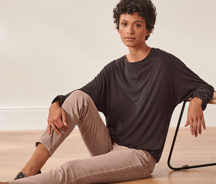 Femme assise par terre à côté d'une chaise en osier, portant un t-shirt manches chauve-souris.