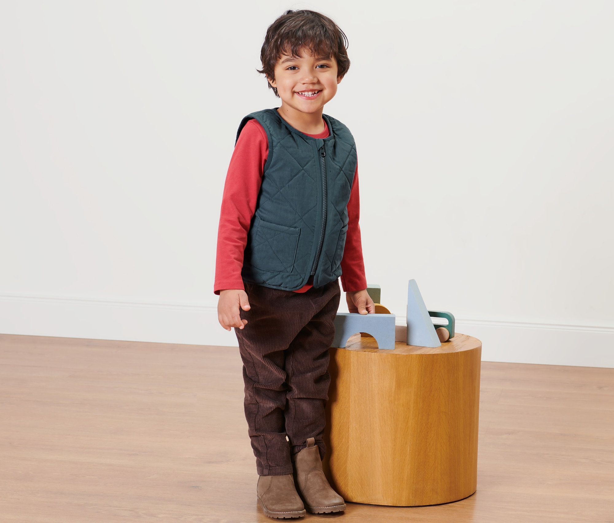 Un garçon se tient à côté d'une table en bois et porte un gilet matelassé réversible pour enfant.