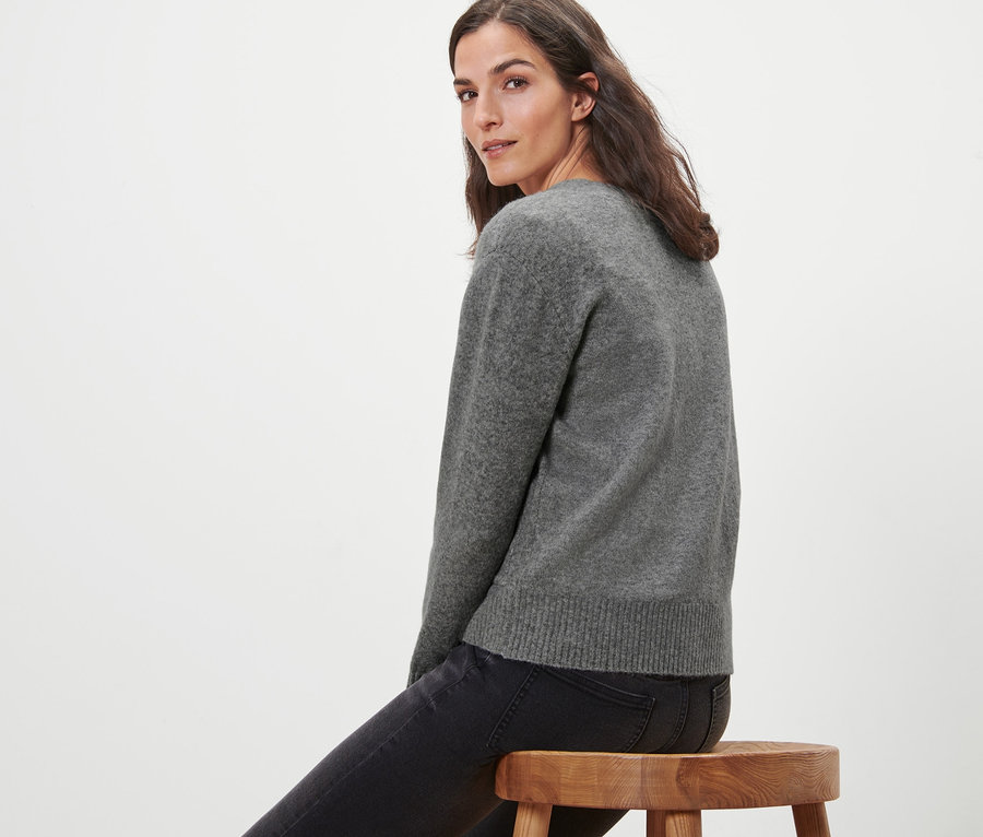 Femme assise sur un tabouret en bois portant une jaquette en maille avec de la laine, gris chiné.