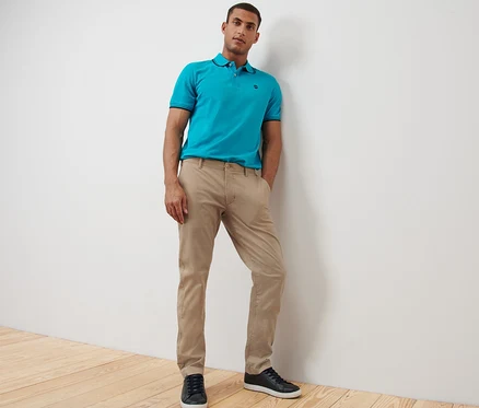 Un homme pose en pantalon chino beige et polo bleu.