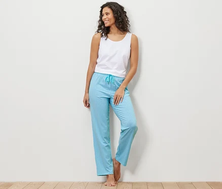 Une femme appuyée contre un mur porte un débardeur blanc et un pantalon de pyjama bleu rayé.