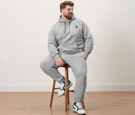 Un homme est assis sur un tabouret et porte un sweat à capuche gris chiné et un pantalon de survêtement.
