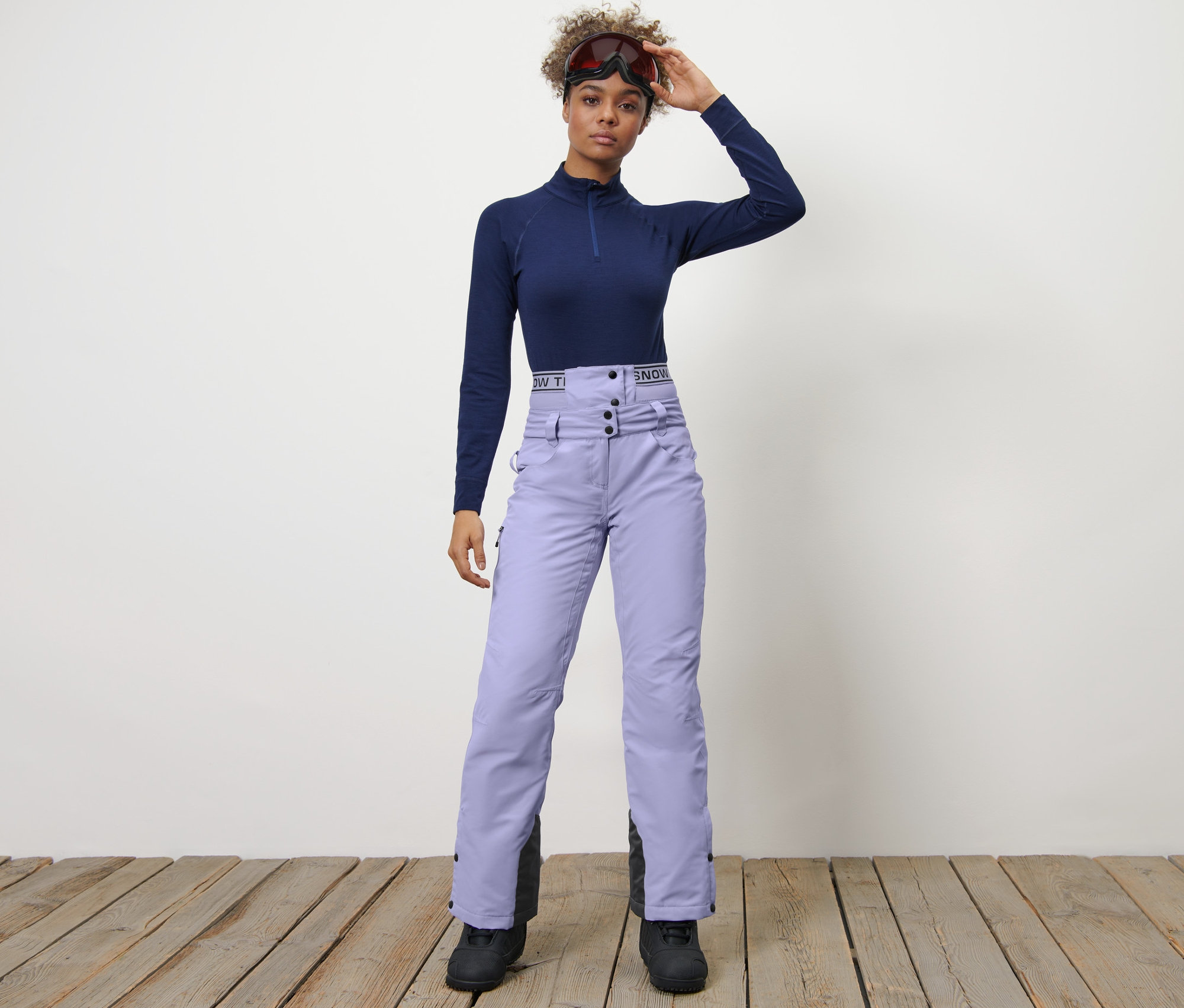 Femme posant en pantalon de ski et de snowboard lilas et haut bleu marine.
