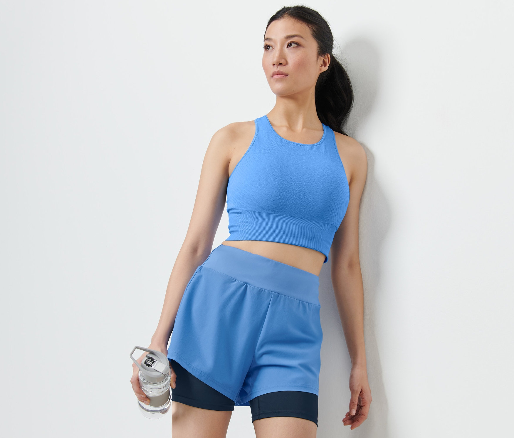 Femme en soutien-gorge de sport bleu et short de sport 2 en 1 tenant une bouteille d'eau.