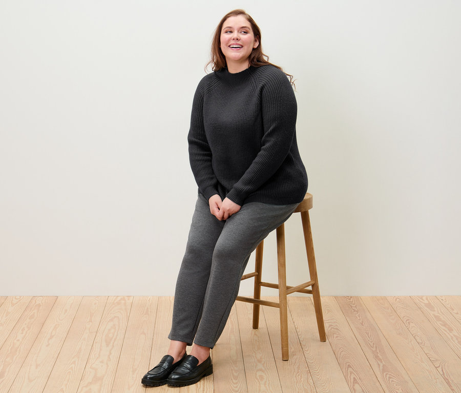 Une femme est assise sur un tabouret et porte un pull noir à col droit en grosse maille et un pantalon de jogging.
