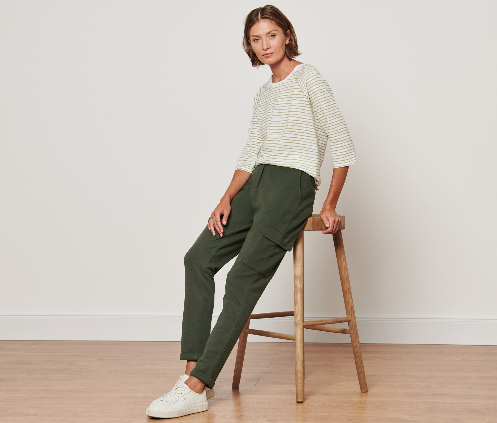 Une femme est assise sur un tabouret en bois et porte un pantalon de jogging vert avec des poches cargo et un haut rayé.