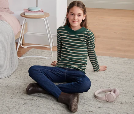 Une fille assise sur un tapis porte un t-shirt côtelé à manches longues rayures vertes, un jean et des bottes.