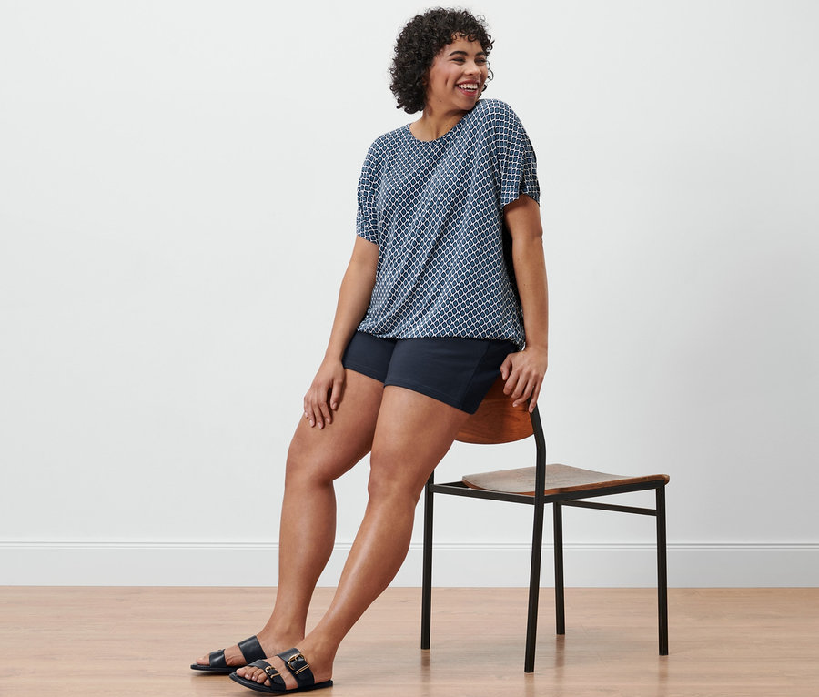 Femme assise de côté sur une chaise portant un chemisier imprimé et un short molletonné bleu marine.