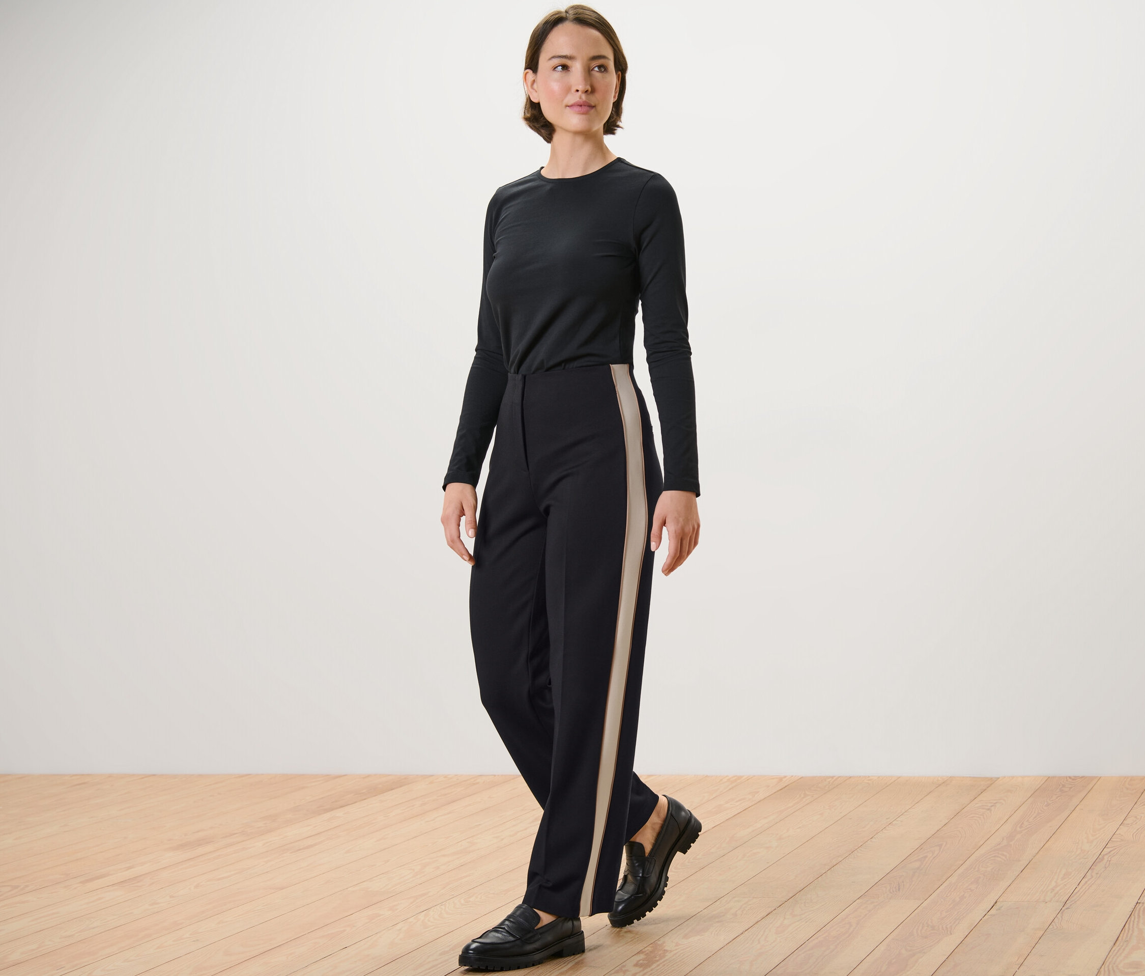 Une femme pose, portant un haut noir à manches longues et un pantalon en jersey noir avec des bandes de galons.