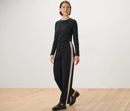 Une femme pose, portant un haut noir à manches longues et un pantalon en jersey noir avec des bandes de galons.