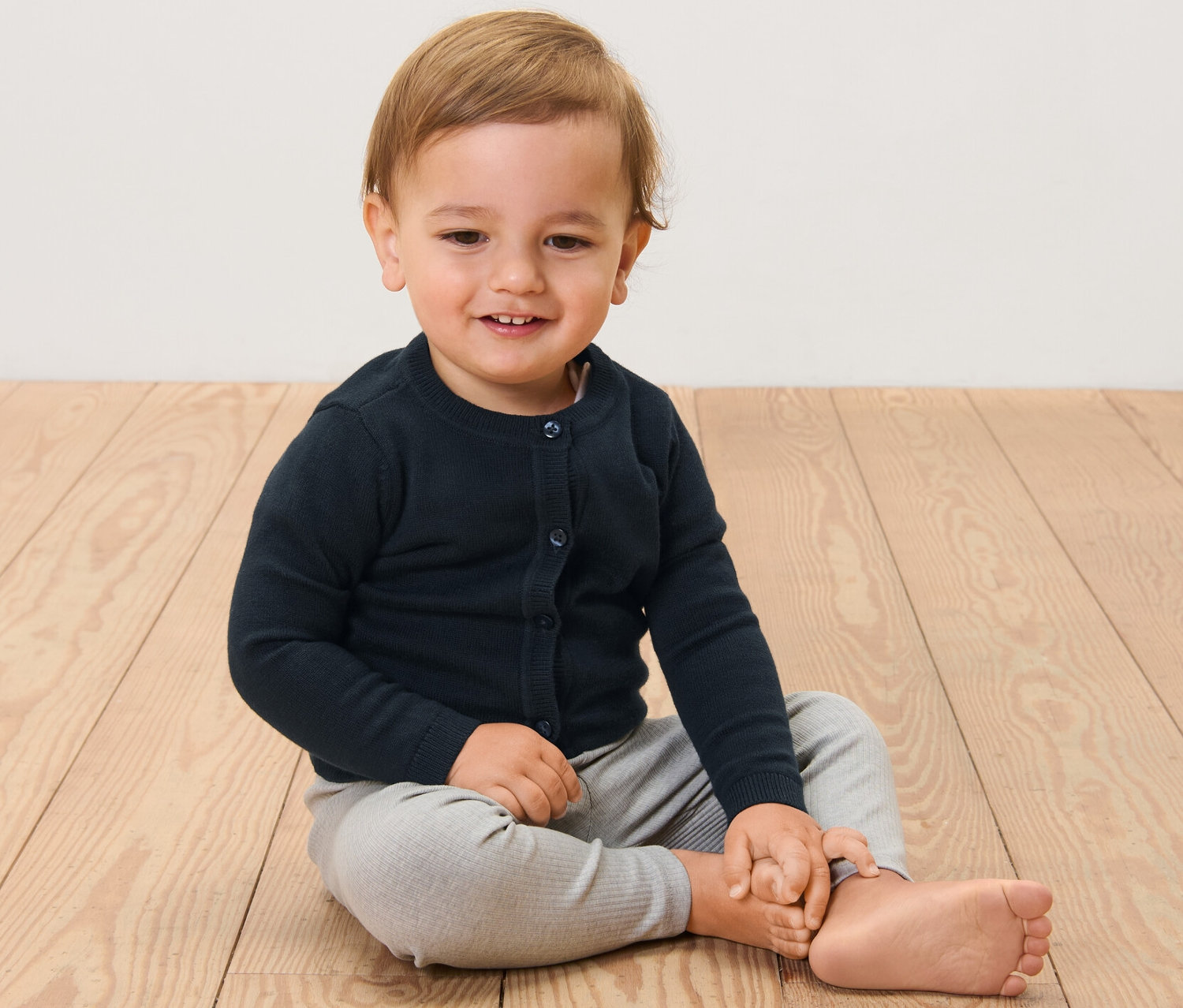 Un enfant est assis sur un plancher en bois et porte une jaquette en maille pour enfant.