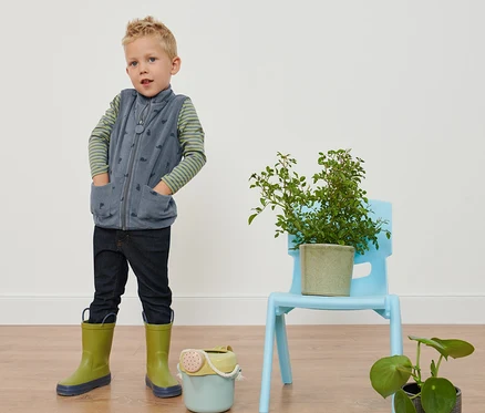 Garçon debout les mains dans les poches portant un gilet en polaire imprimé escargots, un jean et des bottes en caoutchouc.