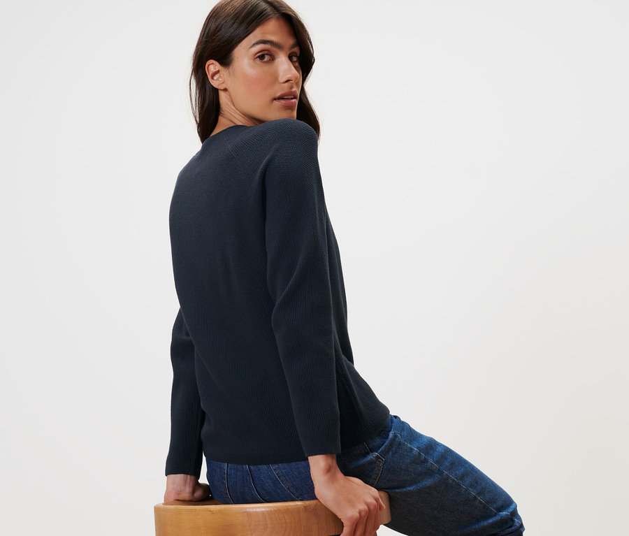 Femme assise sur un tabouret en bois portant un pull en maille bleu foncé.