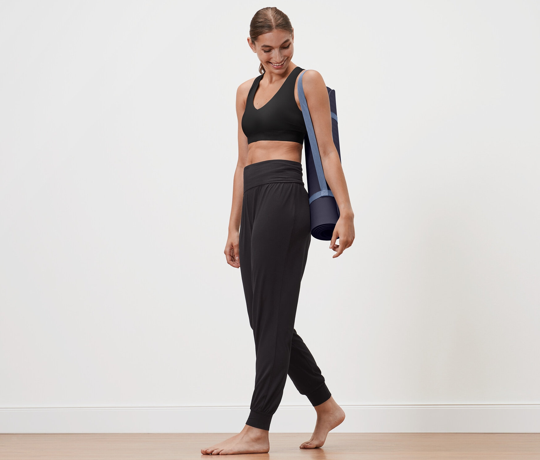 Femme marchant avec un tapis de yoga sur l'épaule. Elle porte un pantalon de yoga.