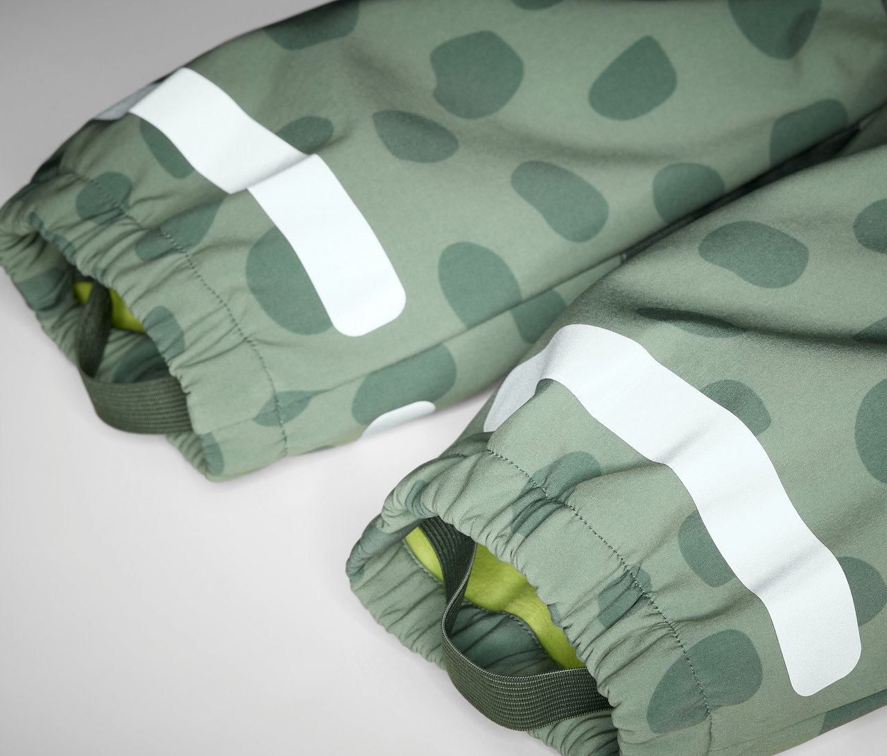 Gros plan sur une combinaison en softshell pour enfant, avec un motif vert olive et une bande réfléchissante blanche.