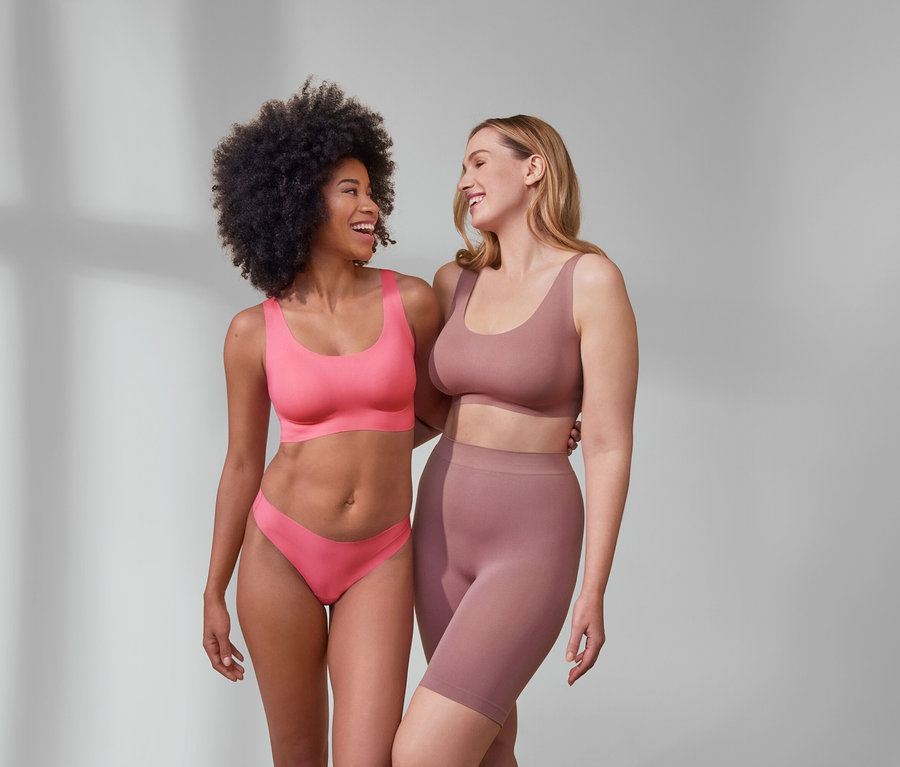Deux femmes sourient et s'embrassent. L'une porte un bustier confortable « bonded », rose vif, et 5 strings Magic Cut. L'autre porte un short sans coutures et un haut.