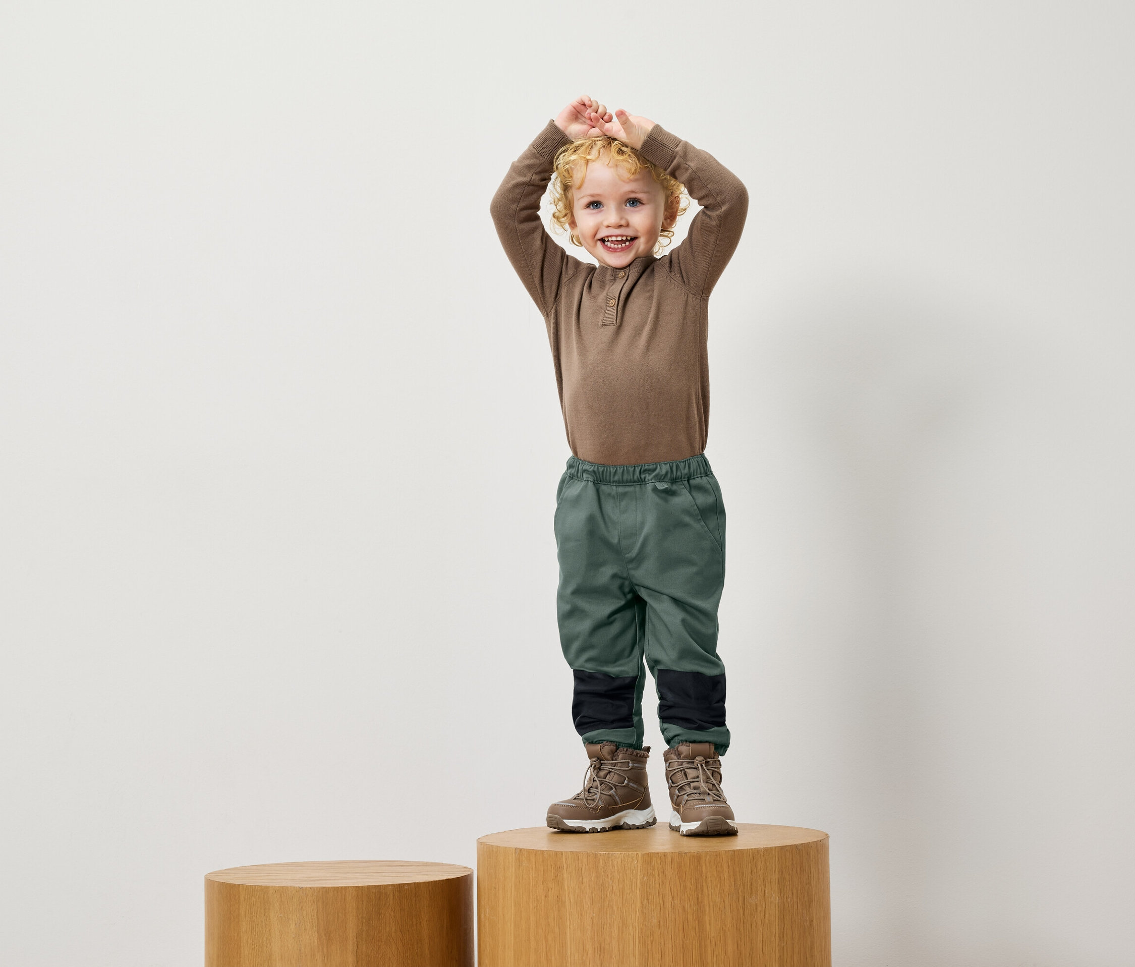 Un garçon aux cheveux bouclés se tient sur un socle en bois, il porte une chemise marron, un pantalon vert et des bottes marron.