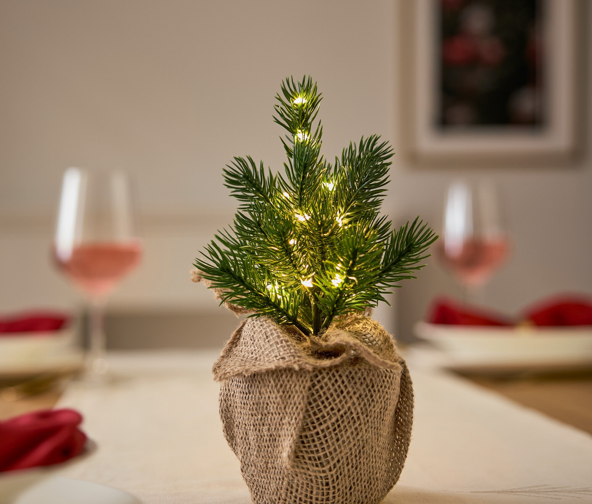 Petit sapin de Noël avec lumières dans un emballage en jute sur une table avec couverts, assiettes et verres.