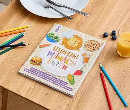 Sur une table, il y a un livre "Restaurant Mitmach Buch" avec des images de nourriture, des crayons de couleur, un verre de jus de fruits et une assiette avec des couverts.