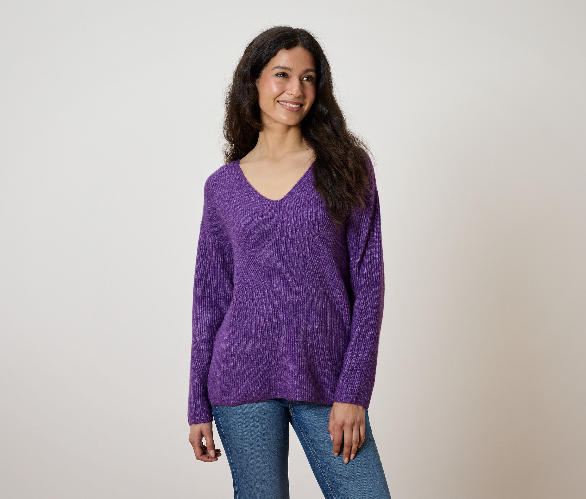 Une femme souriante aux longs cheveux ondulés porte un pull violet à col en V et un jean.