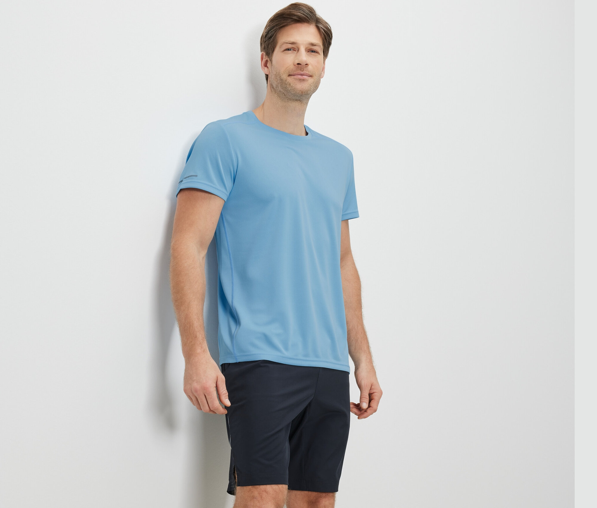 Un homme appuyé contre un mur portant un t-shirt fonctionnel bleu clair.