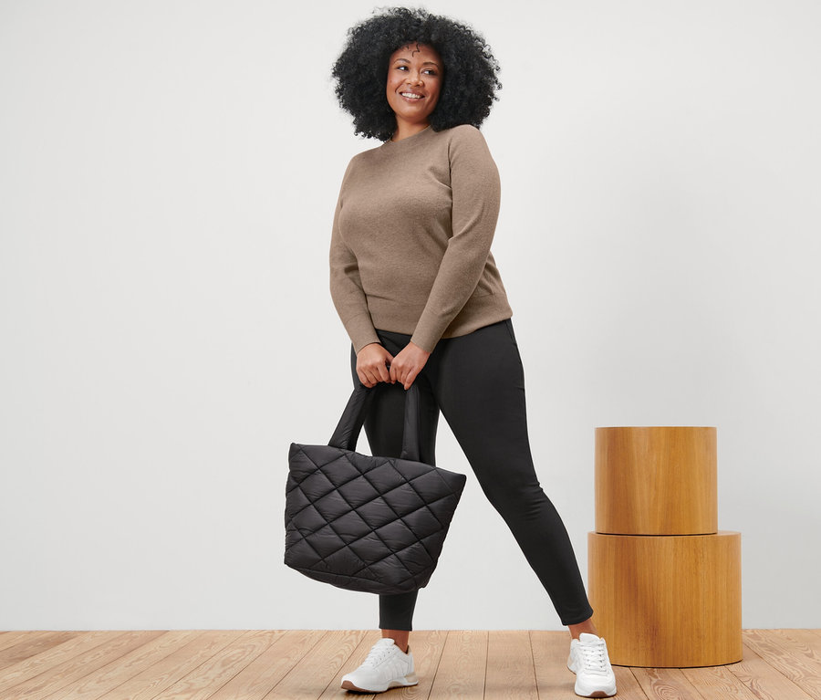 Femme tenant un sac matelassé noir et portant un pull en maille fine beige chiné, un legging noir et des baskets blanches.