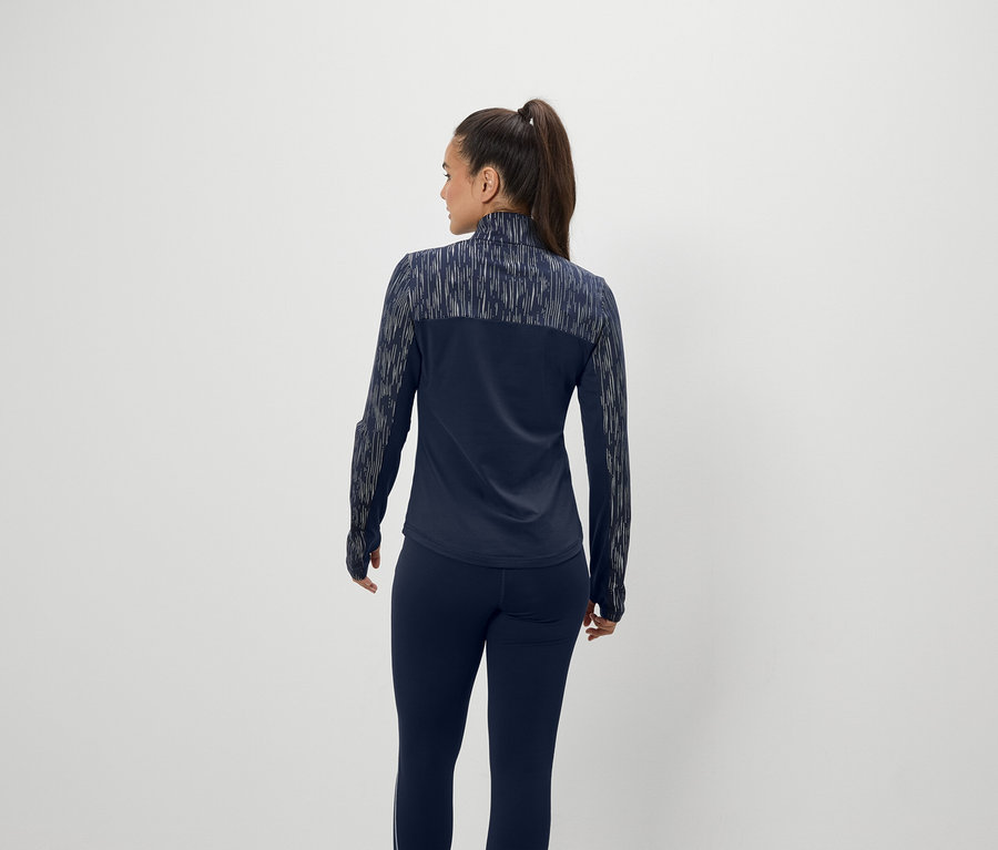 Une femme aux cheveux bruns attachés en queue de cheval se tient dos à la caméra, portant une veste et un pantalon bleu marine.