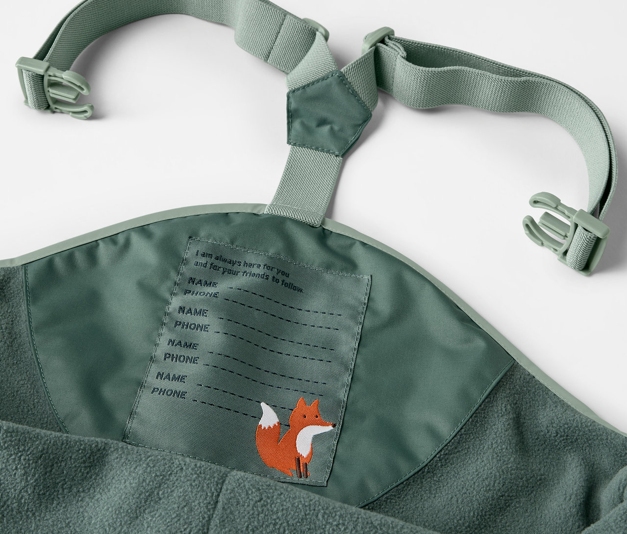 Détail d'un pantalon de pluie thermique pour enfant vert avec bretelles et étiquette de nom avec motif de renard.