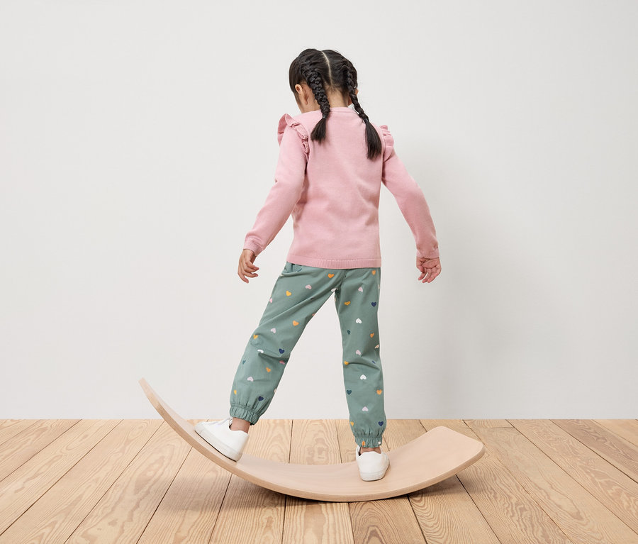Une fillette avec des tresses, vue de dos, en équilibre sur une planche d'équilibre en bois, porte un haut rose et un pantalon vert avec des cœurs.