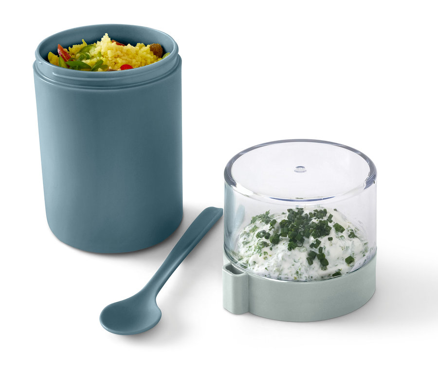 Un récipient de transport pour yaourt vert avec du contenu, à côté d'une cuillère et d'un récipient avec du fromage frais aux herbes.