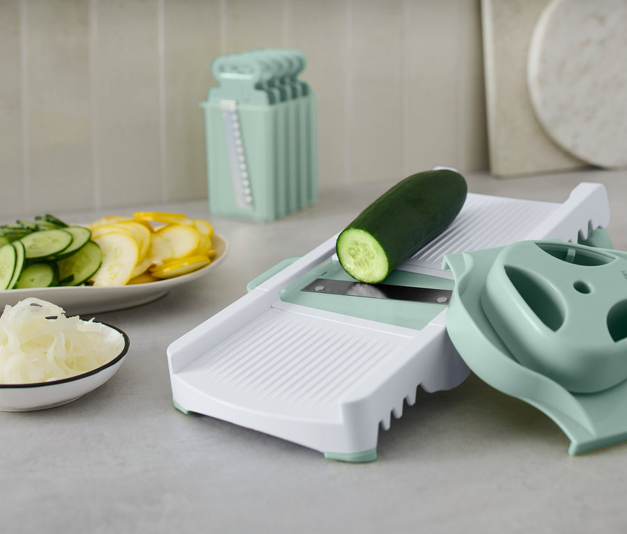Un concombre vert sur une mandoline blanche avec des accessoires. Des concombres et des courges jaunes coupés sont disposés dans une assiette.