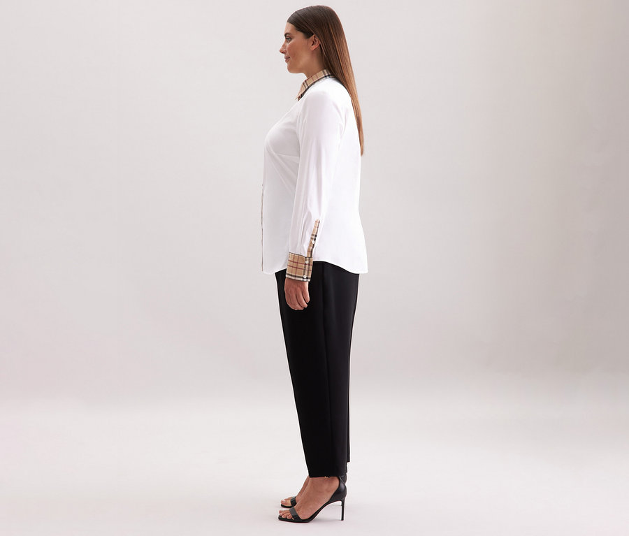 Une femme se tient de profil et porte une chemise en popeline à carreaux Seidensticker blanche, un pantalon noir et des talons hauts noirs.