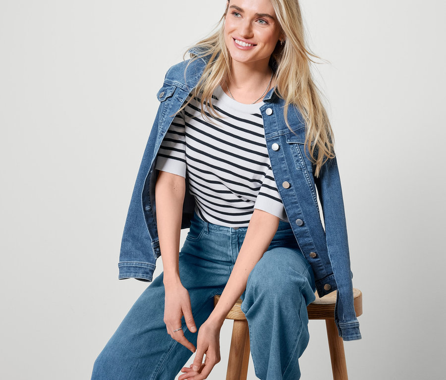 Une femme blonde est assise sur un tabouret en bois et porte une veste en jean, un jean palazzo et un pull en tricot fin rayé.