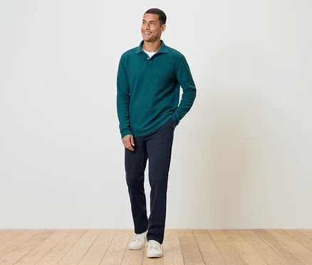 Un homme debout sur un plancher en bois, portant un pantalon bleu marine, un polo à manches longues vert et des baskets blanches.