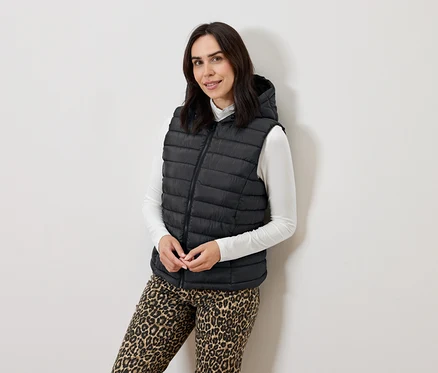 Femme posant dans un gilet noir matelassé à capuche, un pull à col roulé blanc et un pantalon imprimé animal.