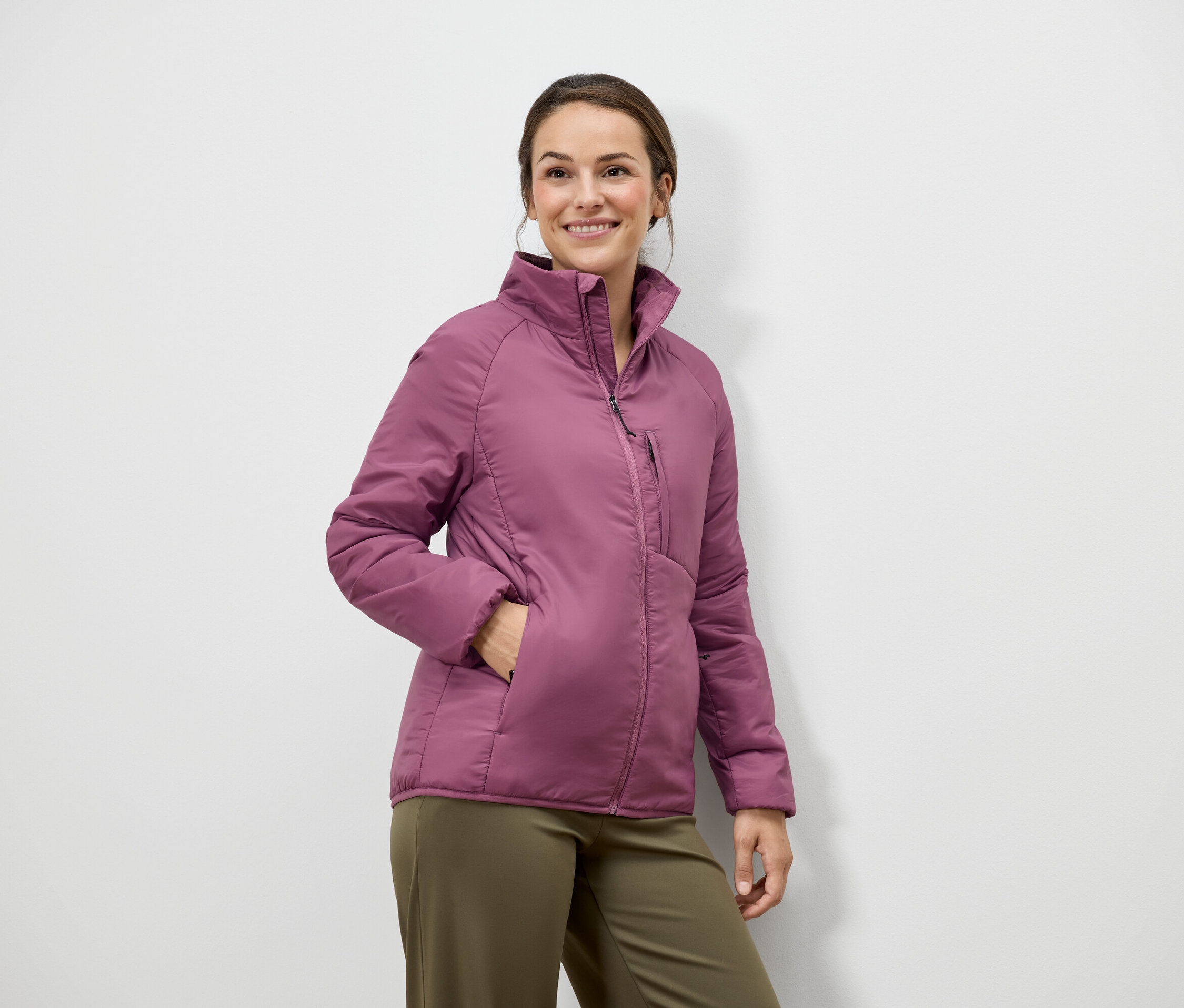 Une femme aux cheveux bruns et un sourire pose en veste rose et pantalon vert devant un mur blanc.