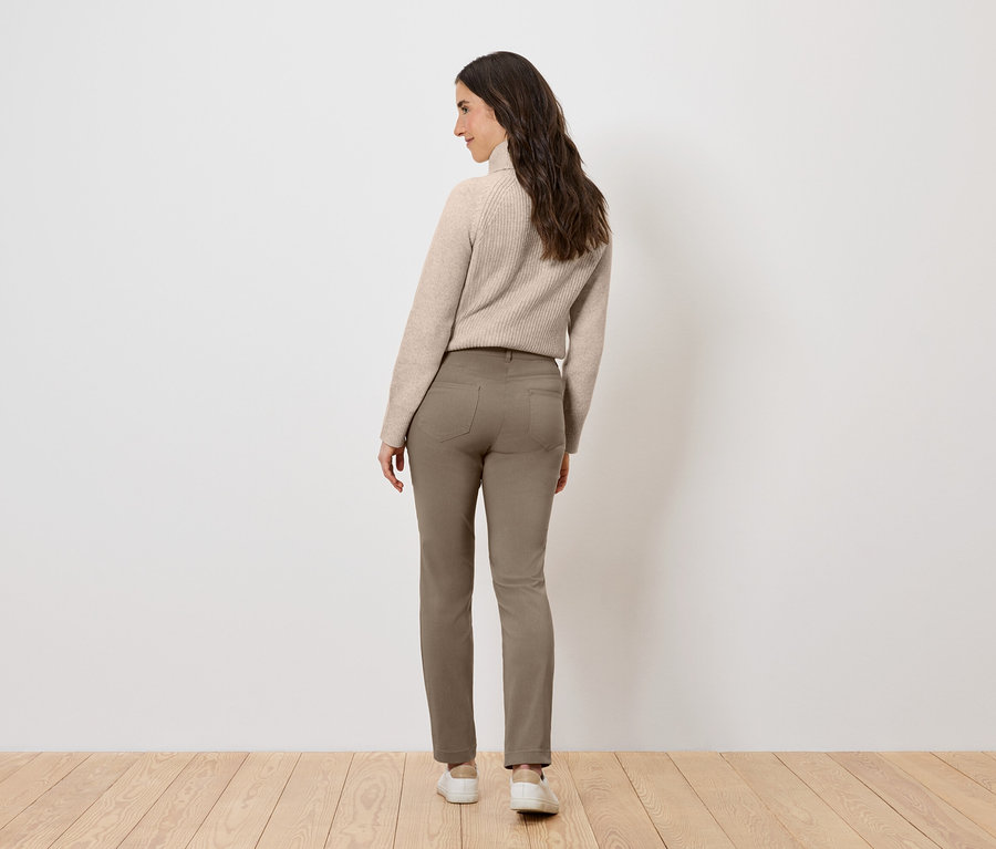 Femme portant un pull beige et un pantalon marron, debout, dos au spectateur.