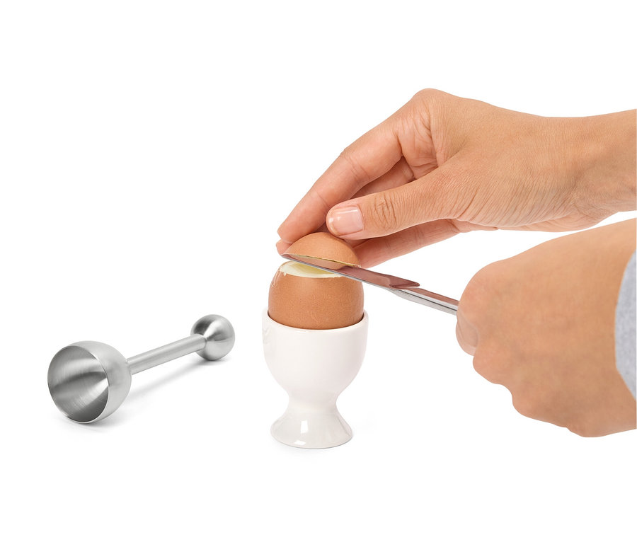 Une main coupe le haut d'un œuf avec un couteau dans un coquetier blanc. Un toque-œuf en acier inoxydable est visible à gauche.