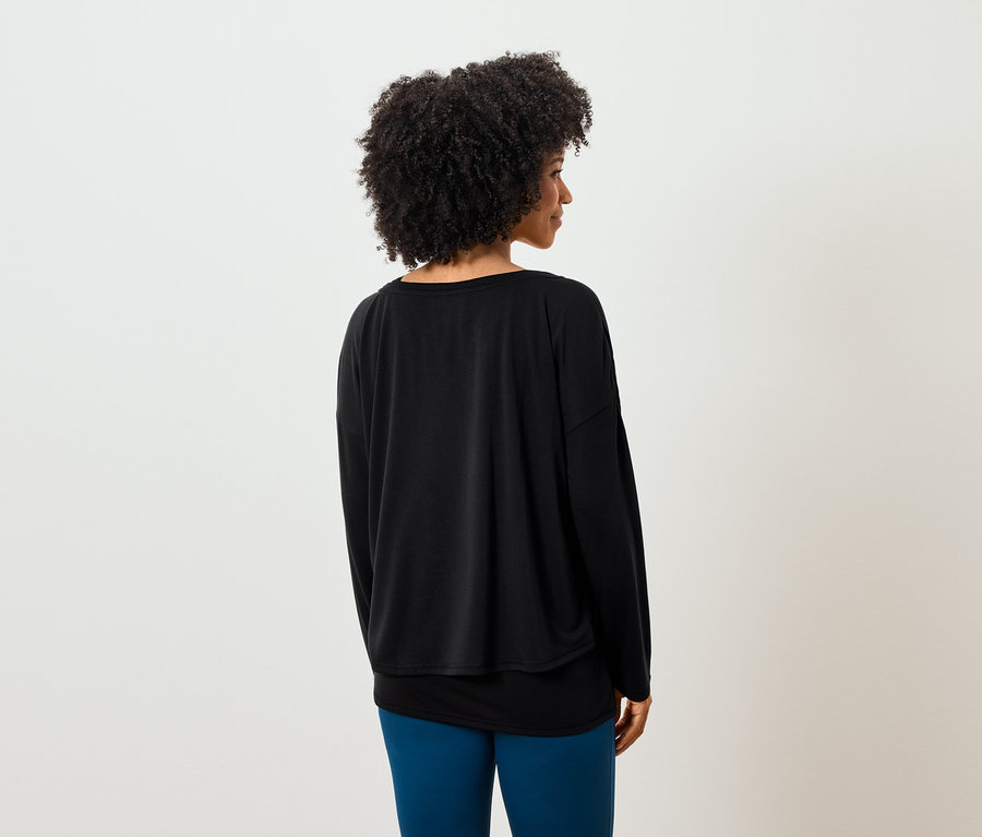 Vue de dos, une femme aux cheveux bouclés porte un t-shirt noir à manches longues et un legging bleu.