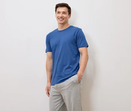 Homme souriant en t-shirt bleu et pantalon de survêtement gris posant sur fond blanc.