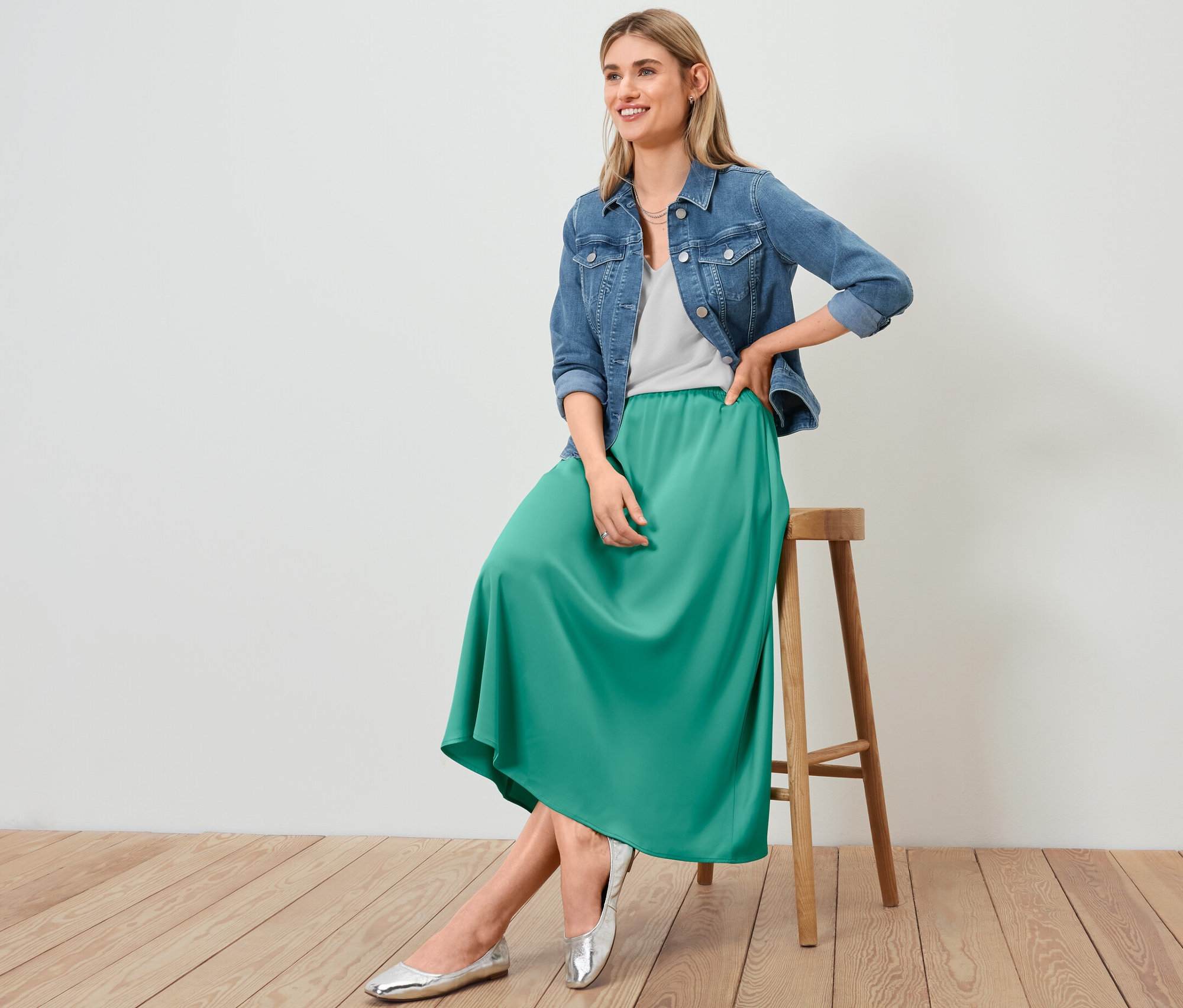 Femme assise sur un tabouret portant une veste en jean, une jupe en satin et une blouse structurée blanche.