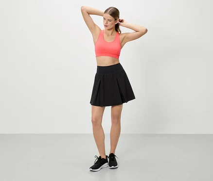 Femme les mains levées portant un haut de sport corail, une jupe noire et des baskets noires.