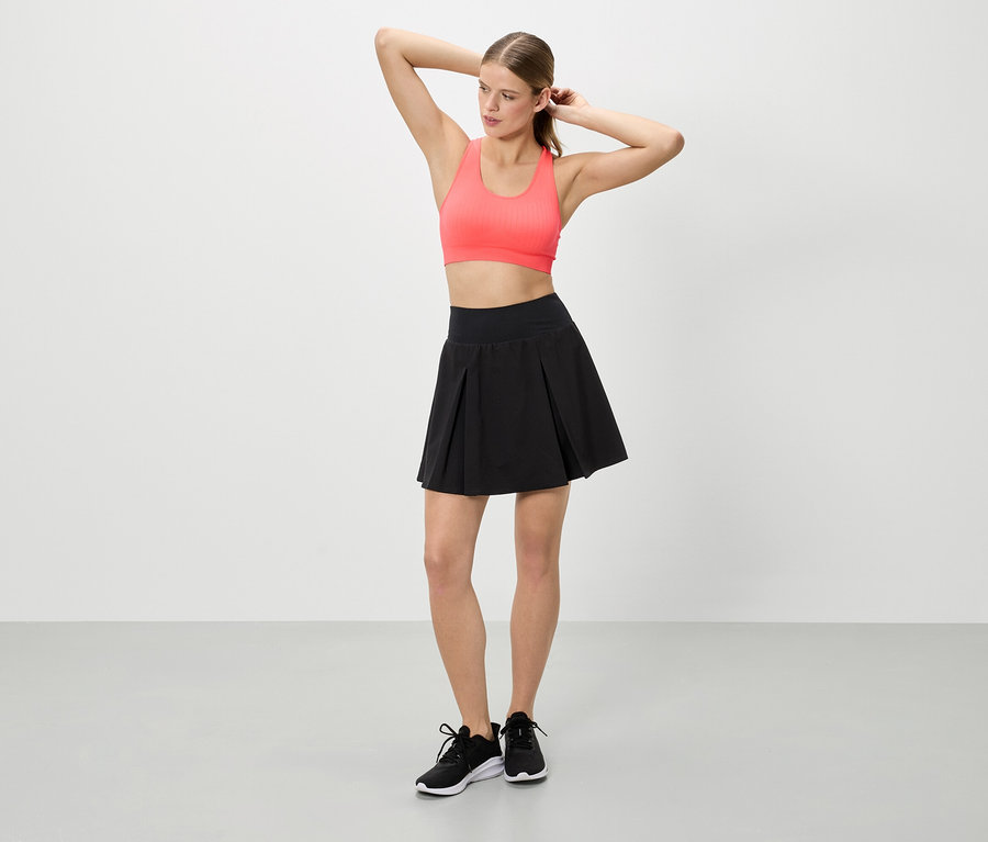 Femme les mains levées portant un haut de sport corail, une jupe noire et des baskets noires.