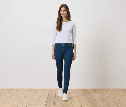 Une femme portant un haut blanc, un jean bleu et des chaussures blanches se tient sur un plancher en bois devant un mur blanc.