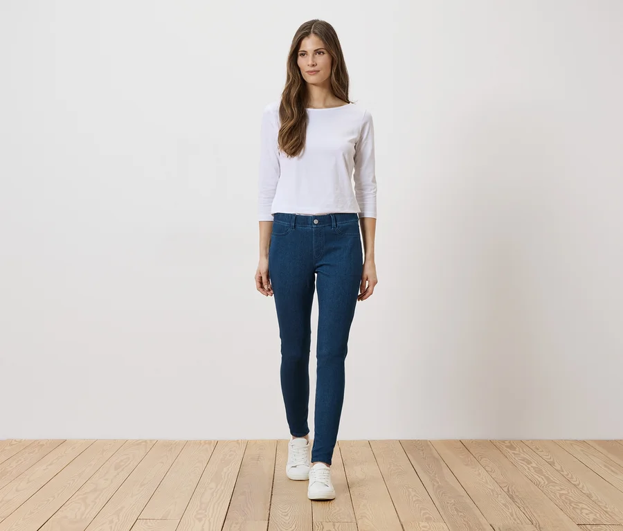 Une femme portant un haut blanc, un jean bleu et des chaussures blanches se tient sur un plancher en bois devant un mur blanc.
