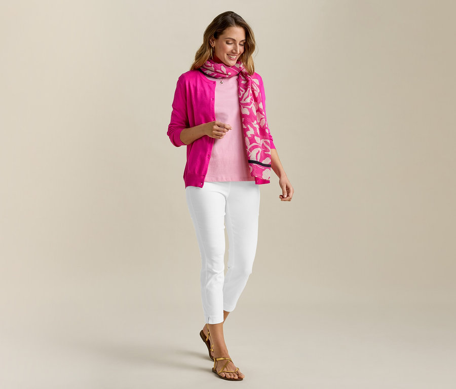 Une femme portant un cardigan en maille fine rose et un pantalon stretch 7/8 blanc. Elle porte un foulard et des sandales.